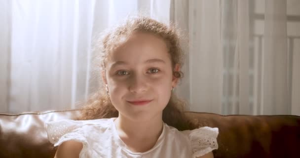 ポートレートカメラを見て笑っている面白い女の子は ソファの上に座っている自宅でソファに座っている かわいい子供ソファに座っている 幸せな子供時代の概念 — ストック動画