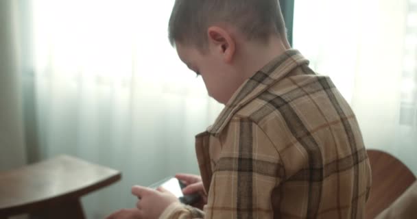 美しい子供の男の子6自宅でスマートフォンでモバイルゲームをプレイ歳 携帯電話で遊んでる可愛い少年 子供はゲームに携帯電話を使う 屋内でビデオゲームをプレイする子供 — ストック動画