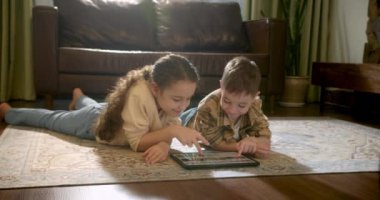 Mutlu iki çocuk, evde sıcak zeminde yatan erkek ve kız kardeş tablet üzerinde oynuyorlar, çocuklar dijital tablet kullanarak videoyu izliyorlar. Çocukluk rüyası, online video oyunu konsepti. küçük çocuklar