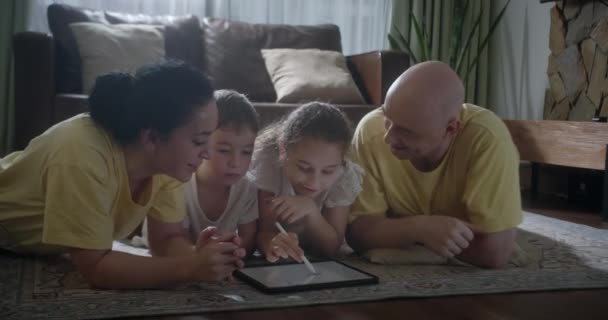 幸福的家庭年轻父母画着色图片用铅笔帮助可爱的孩子女儿享受谈话游戏躺在温暖的地板上在家里 妈妈爸爸和小女孩在客厅休闲的乐趣 — 图库视频影像