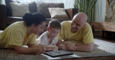 Mutlu bir aile, evde sıcak zeminde yatan genç ebeveynler, anne, baba ve kız boş vakitlerinde oturma odasında bir tablet cihazla evde eğitim görüyorlar. 4K.