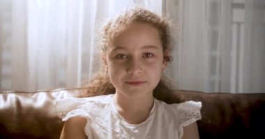 Portre Komik küçük kız kameraya bakıyor evde kanepede oturuyor, güzel yüzlü şirin çocuk kanepede oturuyor. Mutlu bir çocukluk kavramı.