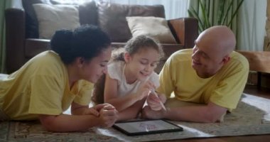 Mutlu aile genç ebeveynleri grafik tablet çizerek tatlı kızın evde sıcak zeminde oturup oyun oynamasını sağlıyorlar anne, baba ve kız boş vakitlerinde oturma odasında eğleniyorlar..