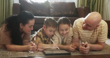 Mutlu bir aile, evde sıcak zeminde yatan genç ebeveynler, anne, baba ve kız boş vakitlerinde oturma odasında bir tablet cihazla evde eğitim görüyorlar. 4K.