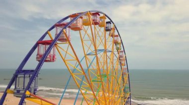 Renkli dönme dolap. Arka planda deniz ve plaj olan bir lunaparkta dönen dönme dolabın havadan görünüşü. Eğlence ve eğlence. Sonsuz döngü. Atlıkarınca eğlendiriyor.