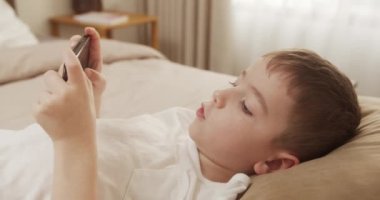 Güzel çocuk. 6 yaşında. Evde akıllı telefondan oyun oynuyor. Cep telefonu çalan sevimli çocuk. Çocuk oyun oynamak için telefonu kullanıyor. İçeride video oyunu oynayan çocuk.