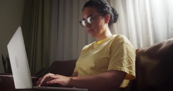 迷人的印度女人坐在沙发上 手里拿着手提电脑 一个人在室内晒太阳 快乐的年轻女性在网上冲浪 在网上冲浪 选择网上购物 — 图库视频影像
