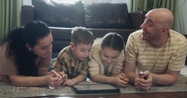 Mutlu bir aile, evde sıcak zeminde yatan genç ebeveynler, anne, baba ve kız boş vakitlerinde oturma odasında bir tablet cihazla evde eğitim görüyorlar. 4K.