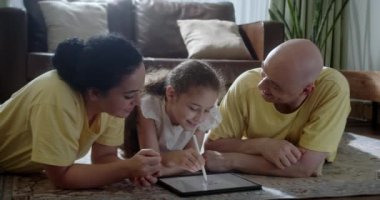 Mutlu bir aile, evde sıcak zeminde yatan genç ebeveynler, anne, baba ve kız boş vakitlerinde oturma odasında bir tablet cihazla evde eğitim görüyorlar. 4K.