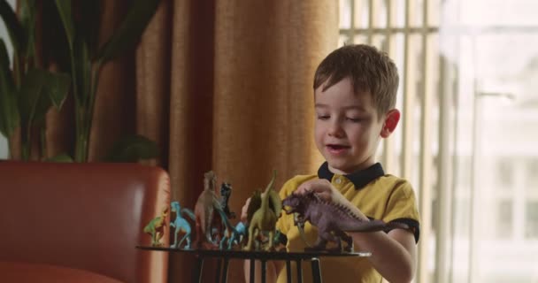 恐竜のおもちゃを演奏するおもちゃと話してかわいい就学前の子供の息子はテーブルの上に座って 幸せな小さな子供は家で楽しんでいる古生物学を説明します — ストック動画