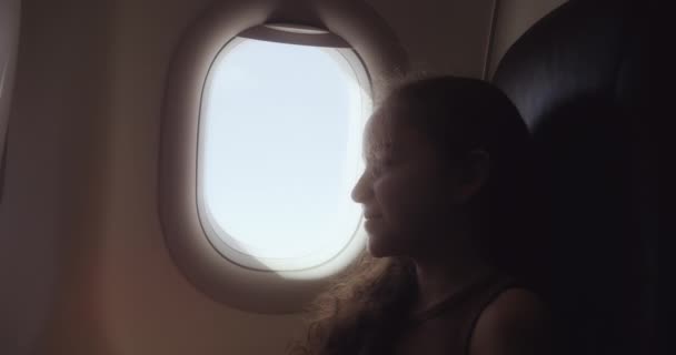 可爱的女孩正坐在飞机的窗前 孩子们在飞行时看着窗外的飞机 迷人的女孩坐着飞机 坐在窗边 坐在航空港孔边的孩子 — 图库视频影像