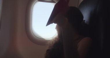 Çocuk uçak penceresinde oturuyor ve uçakta uçarken küçük kağıt uçakla oynuyor. Childs 'ın eli uçak penceresinin arkasında küçük kağıt uçakla. Çocuk 4K' ya bakıyor.