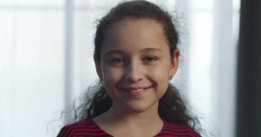 Güzel küçük bir kızın portresi, kırmızı elbiseli gülümseyen bir çocuk, kameraya bakıyor, evdeki kanepede oturuyor, güzel yüzlü şirin bir çocuk kanepede oturuyor. Mutlu çocukluk kavramı. 4K.