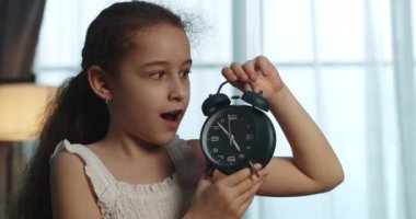 Beyaz elbiseli mutlu küçük bir kız elinde çalar saati tutuyor, çalar saat çalıyor, çocuk endişeli ve neşeli bir yüz ifadesi takınıyor ve saati söylüyor. Eğitici yaşam tarzı. 4K.