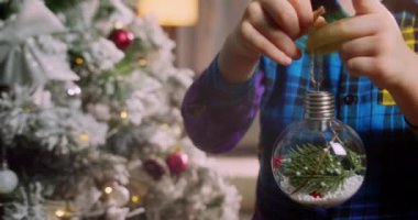 Çocuk elinde Noel ağacı için çok güzel bir Noel balosu tutuyor. Mutlu küçük çocuk Noel ağacını toplarla süslüyor. Ağacın altında bir sürü hediye kutusu var. Çocuk Noel ağacını süslüyor