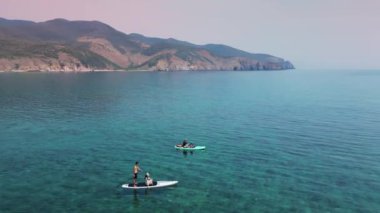 Yaz tatilinde su sporları yapan genç çiftler. Aktif turizm ve seyahat kavramı. Dağlardaki denizde destek için elinde kürek tahtası olan insanların hava görüntüsü. 4K.