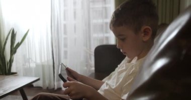 Kanepede tek başına oturan, dijital tablet teknolojisi kullanan meraklı anaokulu çocuğu. Çocuklar teknoloji bağımlılığı konsepti. Home4K 'de küçük çocuk tutucu bilgisayar sörfü internet oyun oyunu.