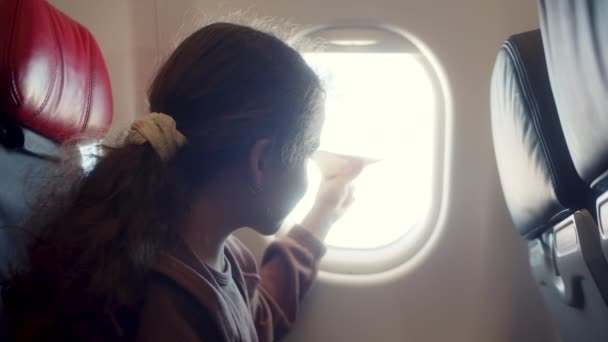 小孩坐在飞机窗边 玩着小纸飞机 在飞机上飞行 在飞机窗口的背景下 孩子们与小纸飞机手牵手 — 图库视频影像