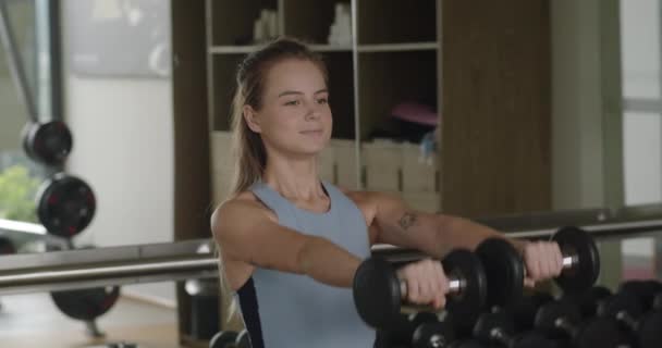 フィットネスルームでスポーツトレーニングを受けるフィットネスガール ジムでボディービルのためのダンベルを持ち上げる女性 少女はスポーツクラブでダンベルを助けて肩のための運動を行います — ストック動画