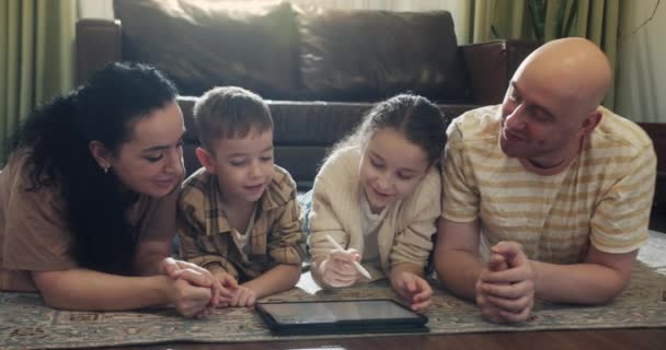 幸せな家族 若い両親は 自宅の暖かい床の上に横たわっています お母さん レジャーでのリビングルームのタブレットデバイス上のお父さんと娘のホームスクーリング — ストック動画