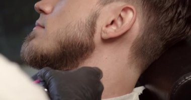 Berber berber dükkanında tıraş makinesiyle sakallı adam buduyor. Saç şekillendirme işlemi. Sakallı bir erkeğin sakalını kesen bir kuaförün yakın çekimi..