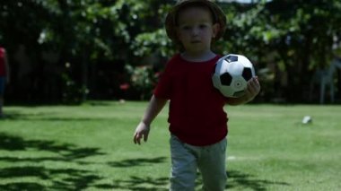Küçük çocuk parkta çimlerin üzerinde futbol oynuyor. Mutlu aile hayali çocuk konsepti. Küçük oğlum parkta sağlıklı bir şekilde top oynuyor, açık hava sporları yapıyor. Çocuk oğluyla oynayıp eğleniyor..