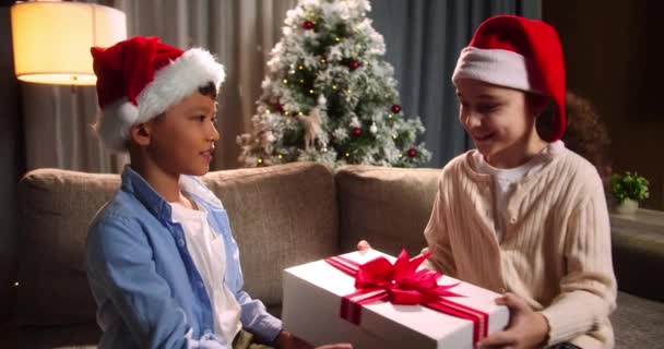 驚きに満ちた幸せなかわいい子供たち 子供たちはクリスマスギフトを交換し 女の子はプレゼントを受け取り ギフトボックスを開きます クリスマスの帽子の中で幸せな子供たちを笑顔肖像 サンタクロース — ストック動画