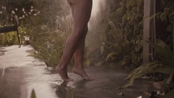 Stock video Young blond woman taking tropical shower.Shower in the private villa. Backside view of woman in swimsuit takes rain shower, tropical greenery on background. Girl washing her head and body.Slow motion