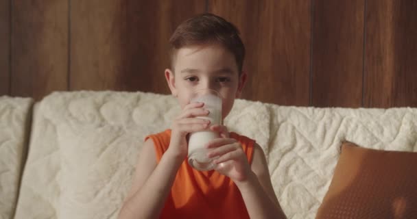 子供のポジティブな子供は テレビを見ながら自宅でソファーに座っている間 ミルクを飲んでいます 朝食を食べる健康的な食べ物の子供 子供の夢の概念 ソファーのリビングルームで息子はヨーグルトミルクを飲みます — ストック動画