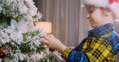 Çocuk elinde Noel ağacı için çok güzel bir Noel balosu tutuyor. Mutlu küçük çocuk Noel ağacını toplarla süslüyor. Ağacın altında bir sürü hediye kutusu var. Çocuk Noel ağacını süslüyor