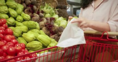 Güzel, genç, esmer, güzel bir kadın marketten yiyecek, domates alıyor. Kız ürünleri, sebzeleri, meyveleri dükkandan seçiyor. Alışveriş, vegan satış konsepti. 4K