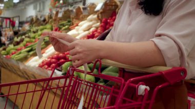 Sebze geçmişi olan bir kadın alışveriş merkezinde alışveriş yaptıktan sonra bakkal arabasındaki Sevgili Miktar faturasını kontrol ederek çekleri kontrol ediyor. Gıda fiyatlarında artış. Bakkal arabasını kontrol eden kadın.