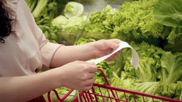 野菜のバックグラウンドに反対する女性は 食料品カートにDear Amount請求書をチェックして モールで食料品の買い物の後に紙チェックをチェックします 食品価格の増加 食料品店のカートを点検する女性 — ストック動画