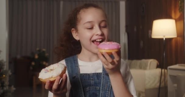 ピンクの釉薬またはホワイトチョコレートで美味しい甘いデザートドーナツを食べるかどうかを決めることができない幸せな11歳の子供の肖像画 甘いペストリーを食べる白人の子供のクローズアップ 4Kについて — ストック動画