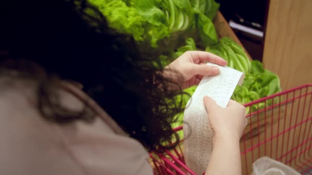 野菜のバックグラウンドに反対する女性は 食料品カートにDear Amount請求書をチェックして モールで食料品の買い物の後に紙チェックをチェックします 食料価格の増加 食料品店をチェックする女性 — ストック動画