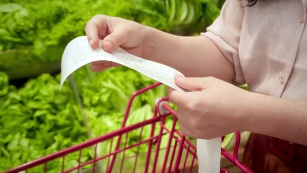 野菜のバックグラウンドに反対する女性は 食料品カートにDear Amount請求書をチェックして モールで食料品の買い物の後に紙チェックをチェックします 食料価格の増加 食料品店をチェックする女性 — ストック動画