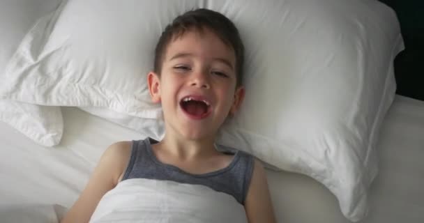 幸せの肖像白いパジャマのかわいい面白い明るい子供の男の子は朝にベッドで目を覚ます 彼の両親を見て笑顔 笑いを伸ばし 毛布の下に再び隠れる — ストック動画