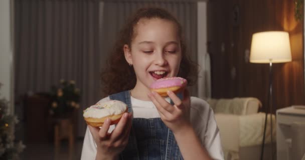 ピンクの釉薬またはホワイトチョコレートで美味しい甘いデザートドーナツを食べるかどうかを決めることができない幸せな11歳の子供の肖像画 甘いペストリーを食べる白人の子供のクローズアップ 4Kについて — ストック動画