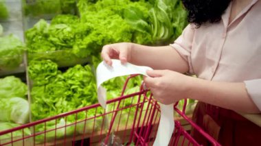 Sebze geçmişi olan bir kadın alışveriş merkezinde alışveriş yaptıktan sonra bakkal arabasındaki Sevgili Miktar faturasını kontrol ederek çekleri kontrol ediyor. Gıda fiyatlarında artış. Kadın markete bakıyor.