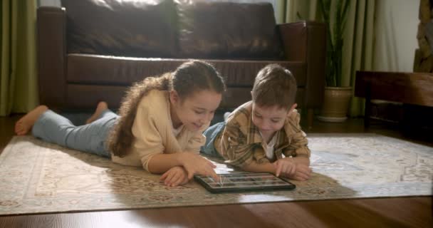 幸せな2人の子供の弟と妹の暖かい床にタブレット上の家庭用ゲームに横たわって 子供たちはデジタルタブレットを使用してビデオを見る 子供の頃の夢 オンラインビデオゲームのコンセプト 幼い子供たちは — ストック動画