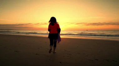 Turuncu bir yaz günbatımında sıcak okyanus suyunda kumların üzerinde koşan bir kadın. Deniz kıyısında, turuncu bir günbatımında, dalgalar boyunca güneşe doğru yürüyen Asyalı ince kadın. 4K 'da yavaş çekim.