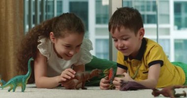 Mutlu küçük çocuklar paleontolojinin evde birlikte eğlenmesini açıklar. Sevgi dolu yetişkin kız kardeş ve tatlı anaokulu erkek kardeşi dinazor oyuncakları oynayarak konuşuyor sıcak zeminde oturuyorlar. 4K.