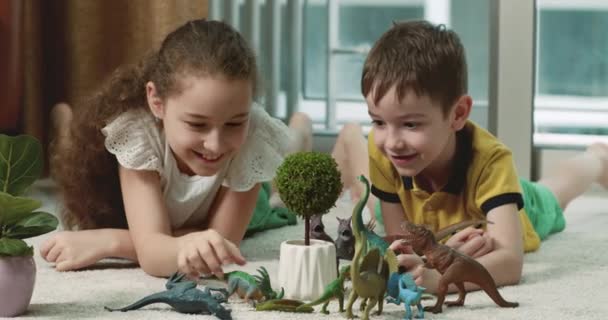 快乐的小孩能解释古生物学家在家里玩得开心的原因 可爱的成年妹妹和可爱的学龄前弟弟在温暖的地板上玩耍恐龙玩具 — 图库视频影像