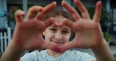 Hoş bir gülümseme takınan mutlu okul kızı kameraya bakıp gülümsüyor ve geceye karşı parmaklarını birbirine dolayıp el yapımı bir kalp sembolü sergiliyor. 4K.