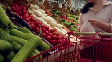 Kız marketten sebze, sebze ve meyve seçiyor. Alışveriş, vejetaryen satış konsepti. Kadın marketten yiyecek, domates alıyor, bir kadın da markette domates sepeti alıyor. 4K.