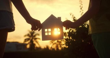 Gün batımında içinde penceresi olan kağıt bir evi kendi elleriyle tutan çocuklar kağıt evin penceresinden güneş ışıldıyor. Evin sembolü, mutluluk. Bir aile için ev inşa etme kavramı