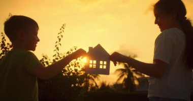 Gün batımında içinde kocaman bir kalp olan kağıttan bir evi ellerinde tutan çocuklar. Güneş pencereden kalplerine doğru parlıyor. Evin sembolü, mutluluk. Bir aile için ev inşa etme kavramı