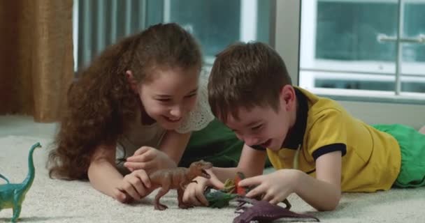 2人の幸せな小さな子供は 自宅のカーペットの上に横たわっている間に床に恐竜と遊びます 自宅でおもちゃと遊ぶコンセプトは 自宅で一緒に楽しむことです おもちゃで遊んでいる子供たち — ストック動画