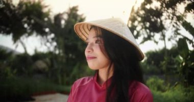 Kameraya bakan mutlu Vietnamlı bir kadının portresi, geleneksel koyu kırmızı bir elbise giyiyor. Genç bir kızın güzel yüzü, tropikal bir geçmişe karşı, Vietnamlı bir...