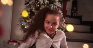 Mutlu çocuk portresi gülümseyen küçük kız kameraya bakarken çok mutlu Noel ağacının yanındaki geleneksel oturma odasında oturuyor. 4K.